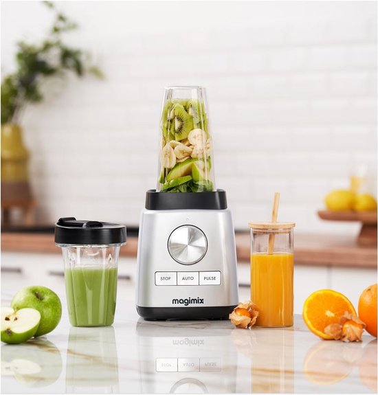 Zilverkleurige Magimix blender met fruit en groenten in de beker, naast een glas groene smoothie en een glas oranje sap.