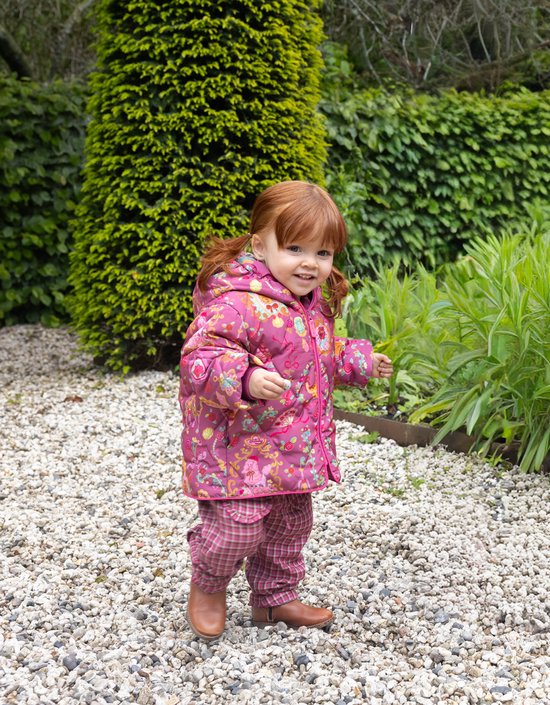 Een jong kind met rood haar, staand in een tuin, gekleed in een roze jas met balletprint en geruite broek.