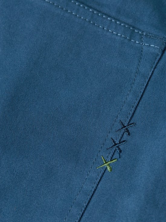 Detailopname van drie gestikte kruisjes, waarvan één in een groene kleur, op de zak van de blauwe broek.