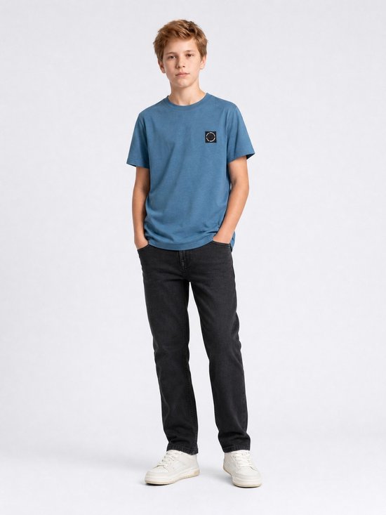 Jongen staat met handen in de zakken, draagt een effen blauw katoenen T-shirt met logo op de borst en zwarte jeans.