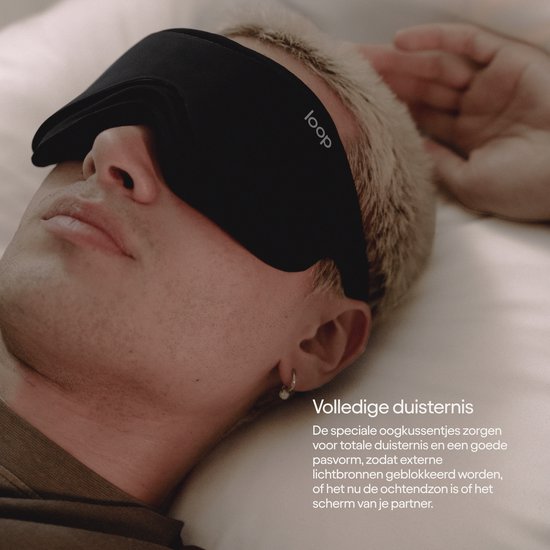 Een man met blond haar slaapt met een zwart Loop-slaapmasker op, met de tekst "Volledige duisternis"