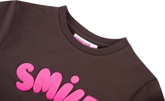 Nekdetail van een bruine sweater met roze "SMILE" print en label.