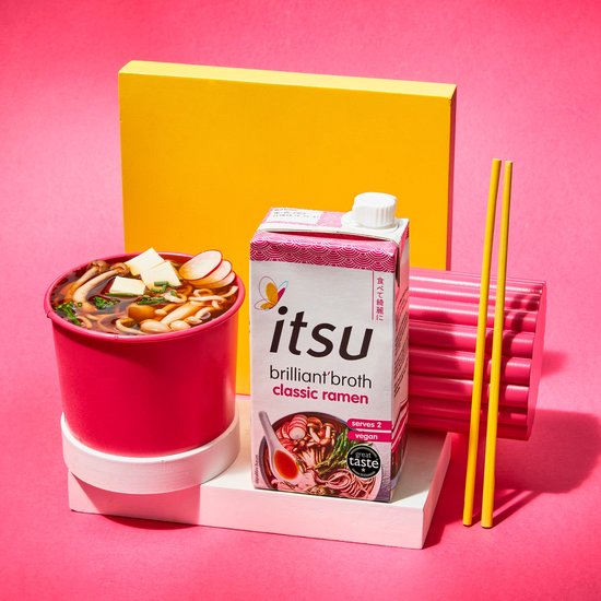 Itsu brilliant broth classic ramen pak naast een rode kom ramen met tofu, radijs en paddestoelen. Gele eetstokjes en roze decoratie ernaast. 'Serves 2 vegan'.