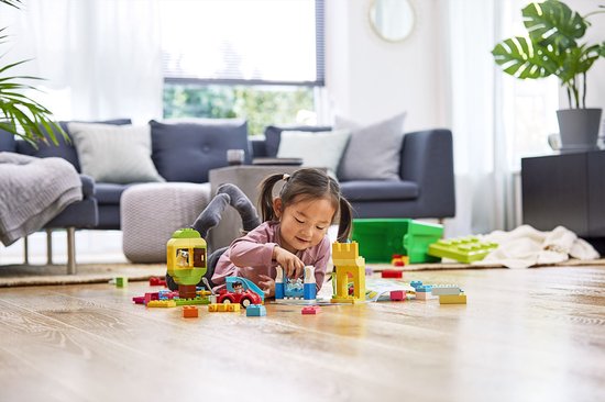 Une petite fille avec des couettes joue sur le sol avec de nombreuses briques LEGO Duplo colorées, dont une voiture rouge et un château.