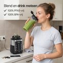 Vrouw drinkt groene smoothie uit een to-go beker naast een zwarte en zilveren blender.
