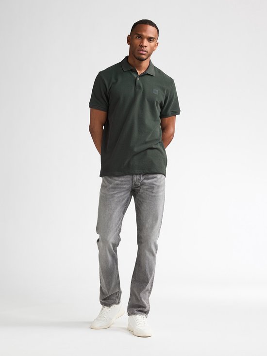 Man staat volledig in donkergroen poloshirt met korte mouwen, grijze jeans en witte sneakers.