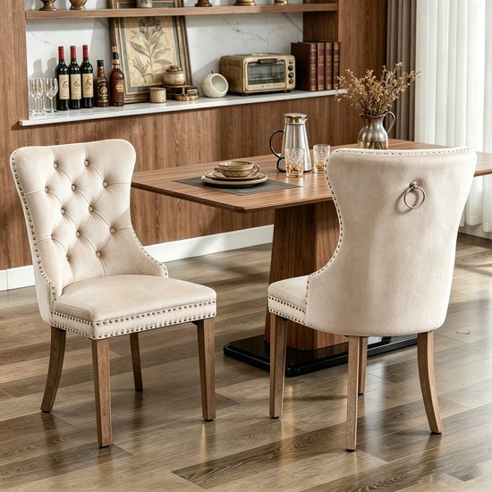 Twee beige fluwelen eetkamerstoelen met knopen, zilveren ringen aan de achterkant en houten poten.