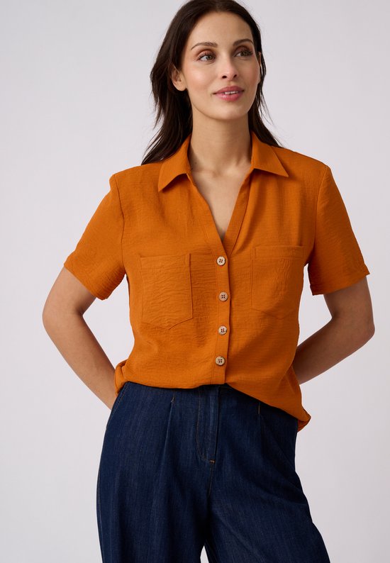 Oranje crêpe blouse met korte mouwen, twee borstzakken en knoopsluiting, gedragen op een donkerblauwe broek.