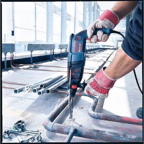 Blauwe en rode Bosch Professional boorhamer met SDS-plus boor, vastgehouden door een persoon met rode werkhandschoenen.