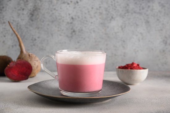 Een glas roze bietendrank met schuim, met een bakje bietepoeder en biet ernaast.