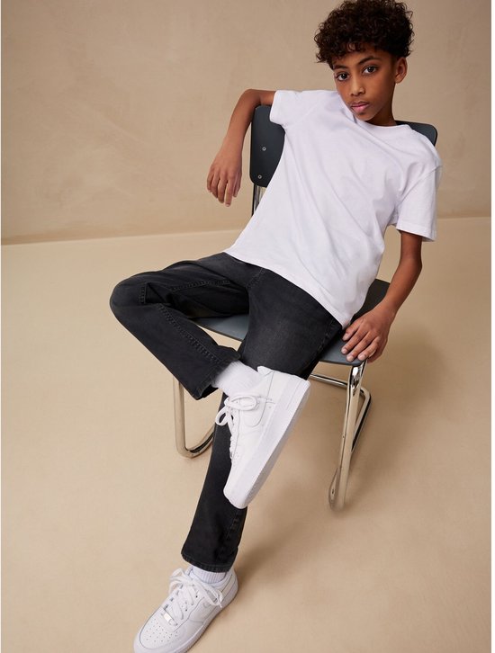 Jongen zittend op een stoel, met witte T-shirt, zwarte spijkerbroek en witte sneakers.