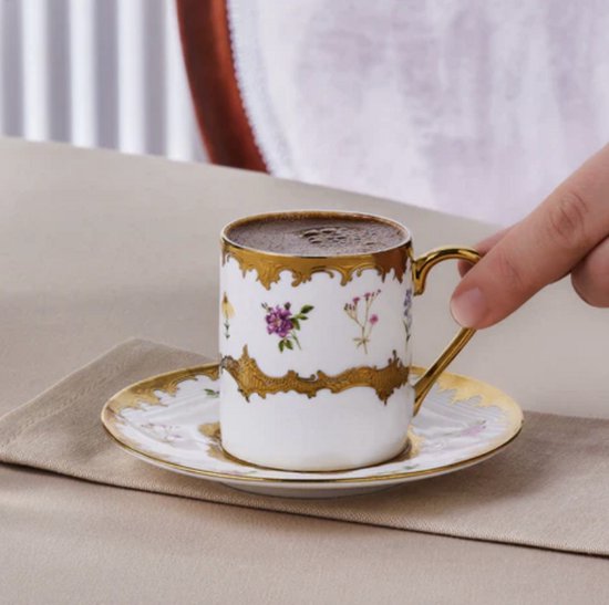 Hand houdt een wit porseleinen koffiekopje vast met gouden rand en bloemen, op een bijpassende schotel.