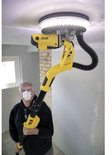 Een man met veiligheidsbril en mondkapje houdt een gele en zwarte telescoopschuurmachine vast die een plafond schuurt.