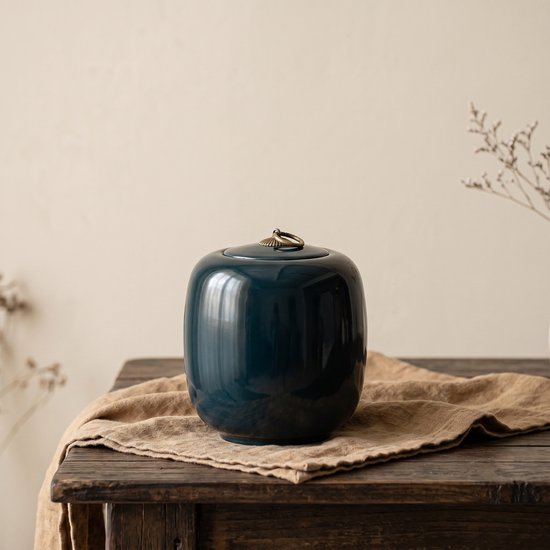 Donkerblauwe, glanzende keramische urn met gouden handvat, staand op een beige doek.