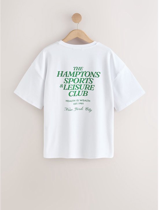 Achterkant van een wit katoenen T-shirt met grote groene tekst 'THE HAMPTONS SPORTS & LEISURE CLUB'.