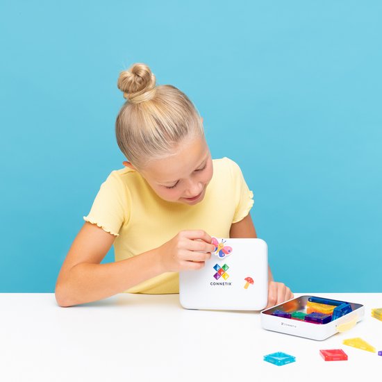 Meisje plakt een sticker van een vlinder op een wit Connetix koffer, met magnetische tegels verspreid.