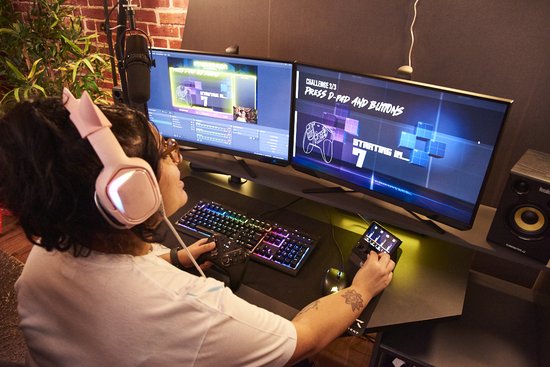 Een streamer met koptelefoon en gamecontroller zit aan een bureau met twee monitoren en de Hercules Stream controller.
