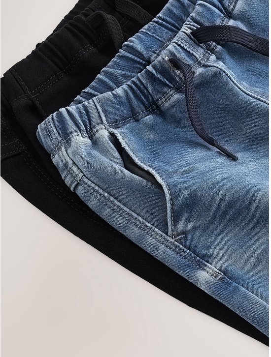 Twee kinderbroeken, een in denimblauw en een in zwart, liggen op elkaar. De taillebanden zijn elastisch met koordjes.