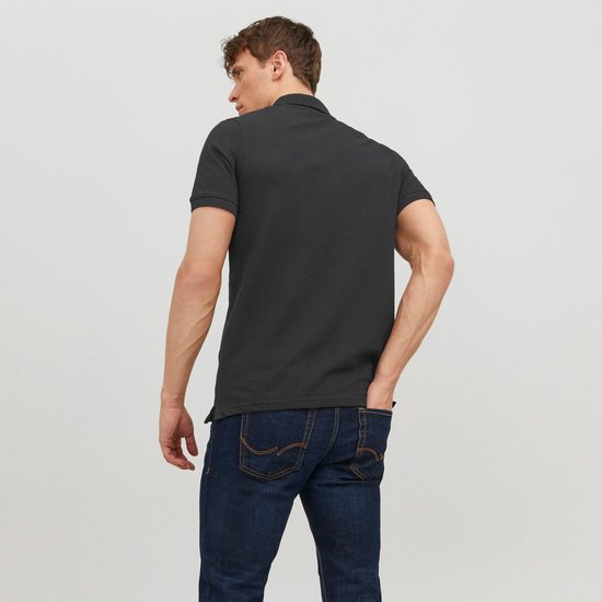 Man met donkergrijze polo en spijkerbroek, staand met de rug naar de camera. Handen in de zakken.