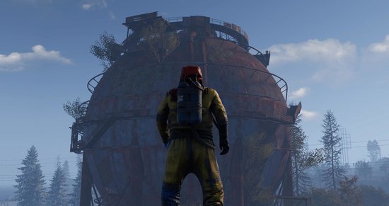 Persoon in geel beschermpak met rode capuchon staat voor een grote, roestige industriële tank.