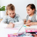 Twee meisjes zitten aan tafel. Eén kleurt in een boek, de ander kijkt toe. Er liggen kleurpotloden.