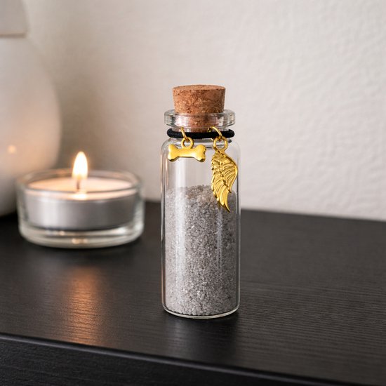 Glazen flesje met as, naast een brandende kaars. Gouden bot- en vleugelhangerjes aan zwart touwtje.