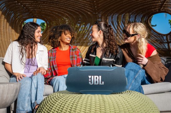 Blauwe JBL Xtreme speaker staat op een groen kussen, gefotografeerd met vier lachende vrouwen op de achtergrond.