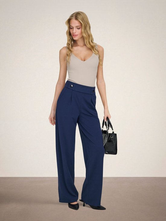 Model draagt donkerblauwe wijde broek met beige top en zwarte tas.