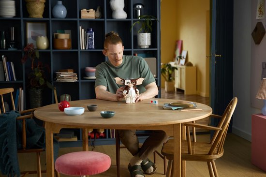 Een man met rood haar, zittend aan een ronde tafel, houdt een LEGO Ideas Gizmo figuur vast. De figuur is gemaakt van bruine en witte blokjes en heeft grote oren.