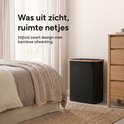 Een zwarte, rechthoekige wasmand met bamboehouten afwerking staat naast een houten ladekast in een slaapkamer met een bed en gordijnen.