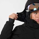 Vrouw trekt capuchon van zwarte ski-jas omhoog, met ski-bril zichtbaar.