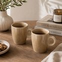 Twee beige mokken met handvat, gevuld met koffie, op een houten tafel.