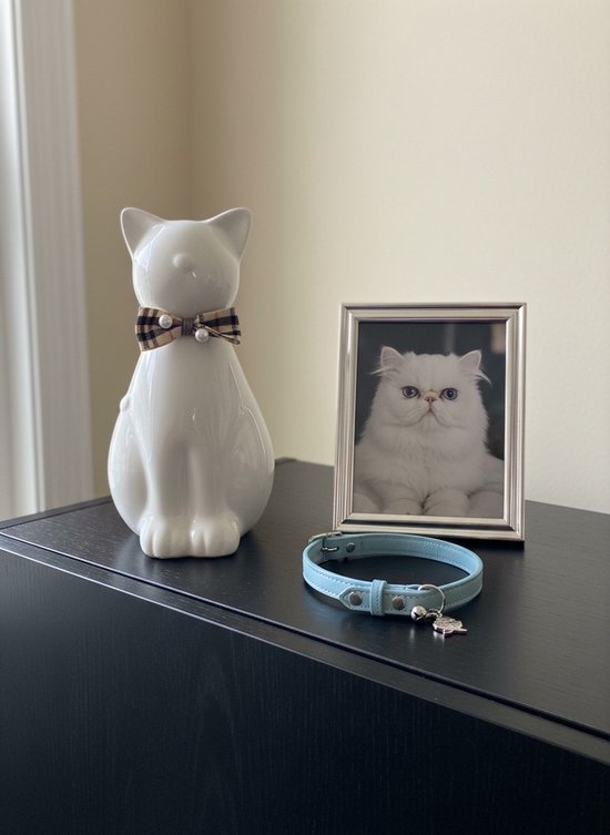 Witte keramische kattenurn met ruitjesstrik en parels, naast een fotolijstje van een kat en een blauw halsbandje.