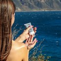 Een vrouw met lange bruine haren houdt een tube Altruist Face Fluid SPF 30 boven haar hand bij een blauwe zee.