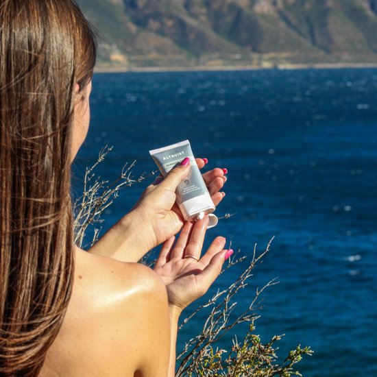 Een vrouw met lange bruine haren houdt een tube Altruist Face Fluid SPF 30 boven haar hand bij een blauwe zee.
