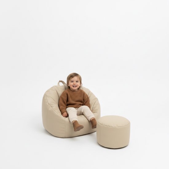 Een jong kind in een bruine trui en beige broek zit in een beige zitzak met plooien, naast een beige poef.