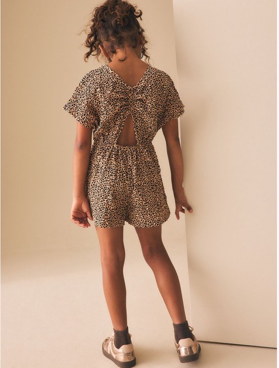 Meisje met luipaardprint playsuit, korte mouwen, open rug met strikdetail en gouden sneakers.