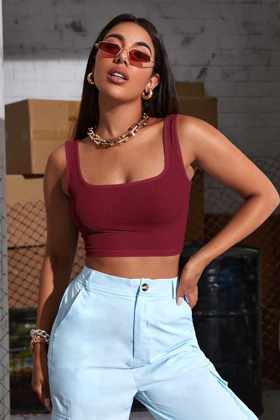 Model in bordeauxrode crop top met vierkante hals en lichtblauwe broek. Gouden ketting en armband.