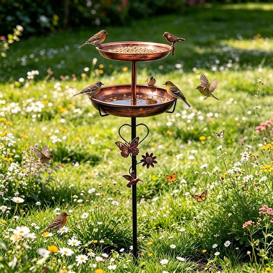 Koperkleurig dubbel vogelbad en voederstation op voet, versierd met vlinders en bloemen, in een weelderige bloementuin.