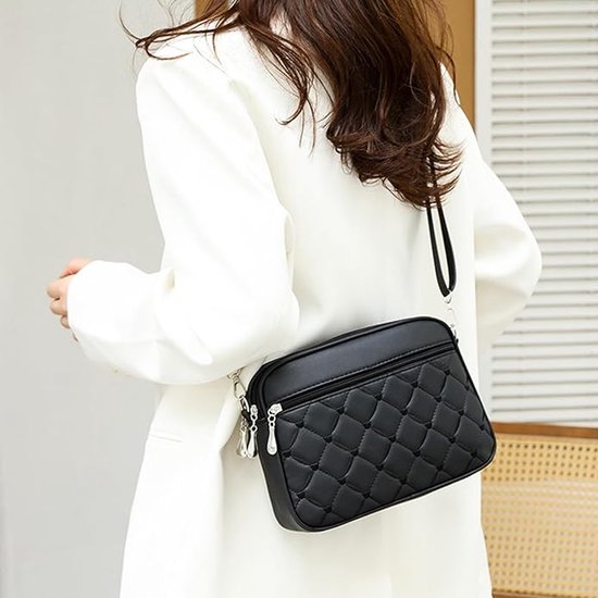 Zwarte gewatteerde crossbody tas met ritsvak aan de voorzijde, gedragen op de schouder door een vrouw in een wit jasje.