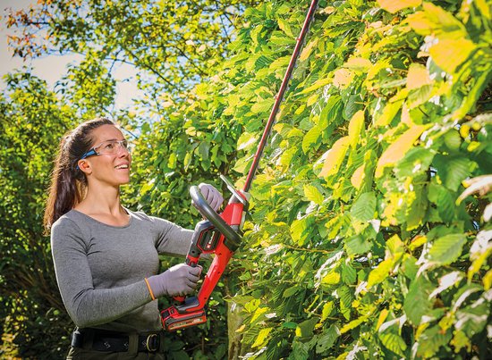Een vrouw met veiligheidsbril snoeit een groene heg met een rode Einhell heggenschaar.
