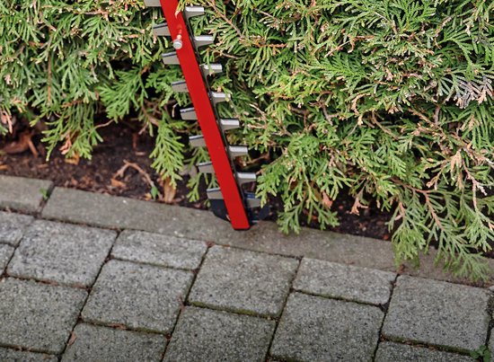 Einhell heggenschaar met rode behuizing en zilveren messen die in een groene coniferenhaag snijdt.