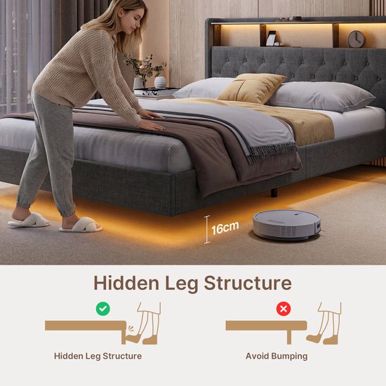 Une femme pousse un aspirateur robot sous un lit rembourré gris foncé avec une structure de pieds bas et cachés.