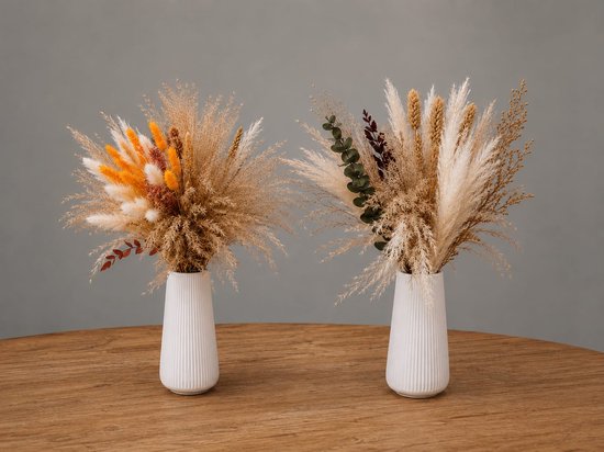Twee droogboeketten van pampasgras met oranje accenten en eucalyptus in witte vazen op een houten tafel.