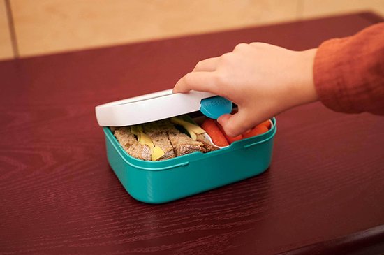Un enfant ferme une boîte à lunch turquoise remplie de sandwichs et de carottes.