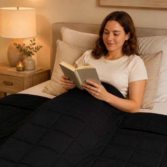 Vrouw leest een boek in bed onder een zwarte, gewatteerde verzwaringsdeken.