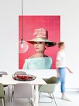 Audrey Hepburn portant un grand chapeau de paille orné d'un nœud rose, un haut pâle, sur fond rose vif. Une femme passe au second plan.