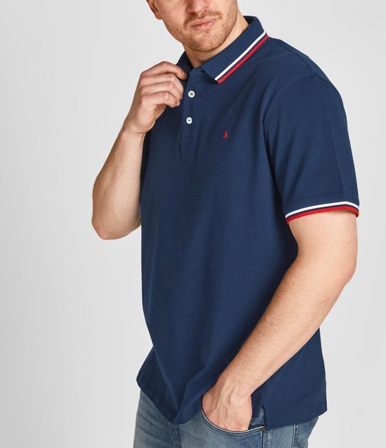 Donkerblauwe polo met rood-wit gestreepte kraag en mouwboorden. Rood logo op borst. Man met hand in broekzak.