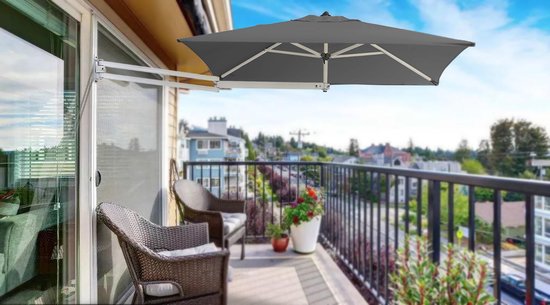 Donkergrijze rechthoekige muurparasol, geopend, boven een balkon met twee stoelen en uitzicht op huizen.