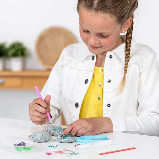Meisje met blonde vlecht versiert een steen met strass steentjes met een roze applicatiepen.